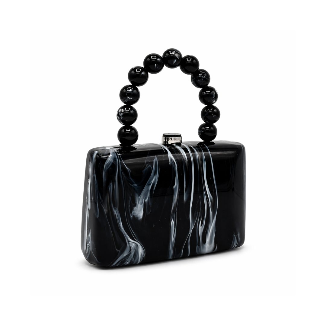 L’APOGÉE Cruella structured acrylic clutch featuring glossy black marbled textures, a rectangular silhouette, and a sculptural black beaded handle designed for edgy evening styling and modern monochrome fashion looks.