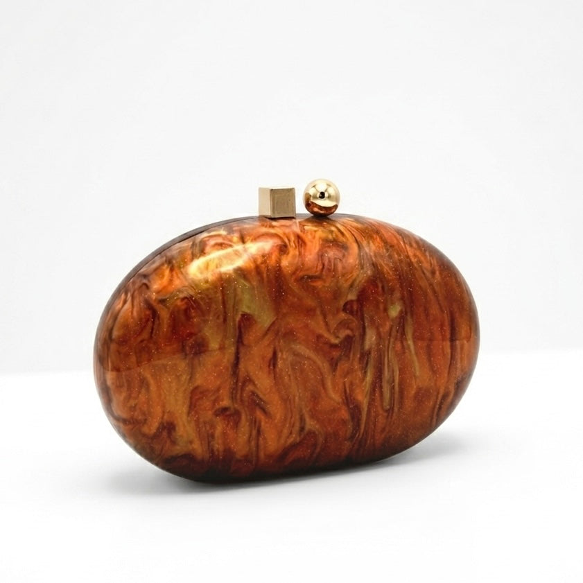 L’APOGÉE Ombrée oval acrylic clutch featuring rich caramel brown and amber marbled textures, a glossy sculptural silhouette, and gold metallic clasp detailing designed for elegant evening styling and luxury fashion moments.