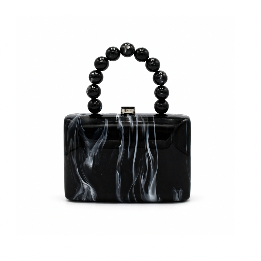 L’APOGÉE Cruella structured acrylic clutch featuring glossy black marbled textures, a rectangular silhouette, and a sculptural black beaded handle designed for edgy evening styling and modern monochrome fashion looks.
