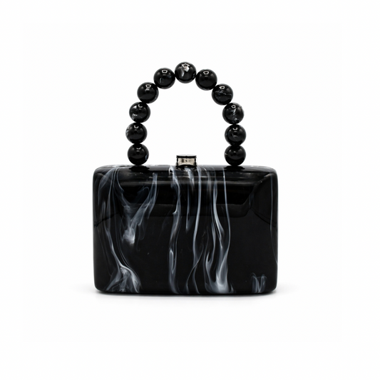 L’APOGÉE Cruella structured acrylic clutch featuring glossy black marbled textures, a rectangular silhouette, and a sculptural black beaded handle designed for edgy evening styling and modern monochrome fashion looks.